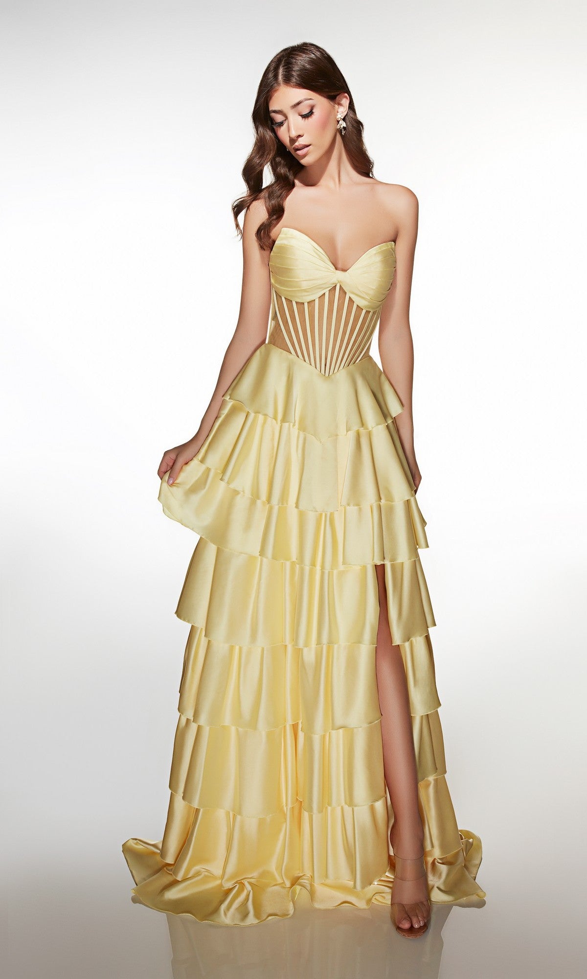 A woman wears the Long Prom Dress: Alyce 62056—a strapless satin gown with a sweetheart neckline, yellow tiered ruffle A-line skirt, corset-style bodice, and high slit—standing against a plain white background.